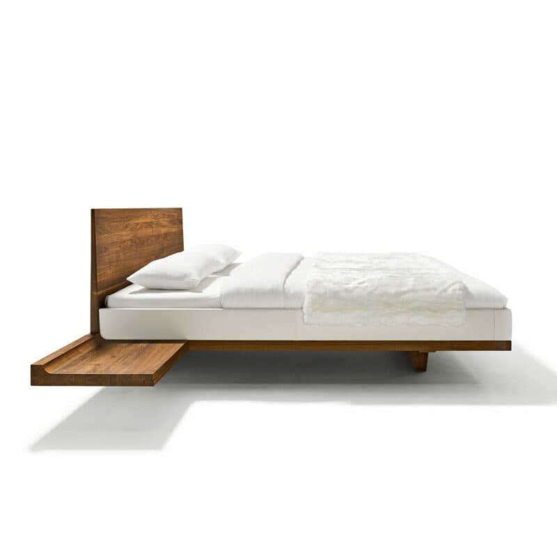 Team 7 „Riletto“ Bett in Nussbaum mit Lederbezug Origon in Weiß inklusive Konsole in seitlicher Ansicht.
